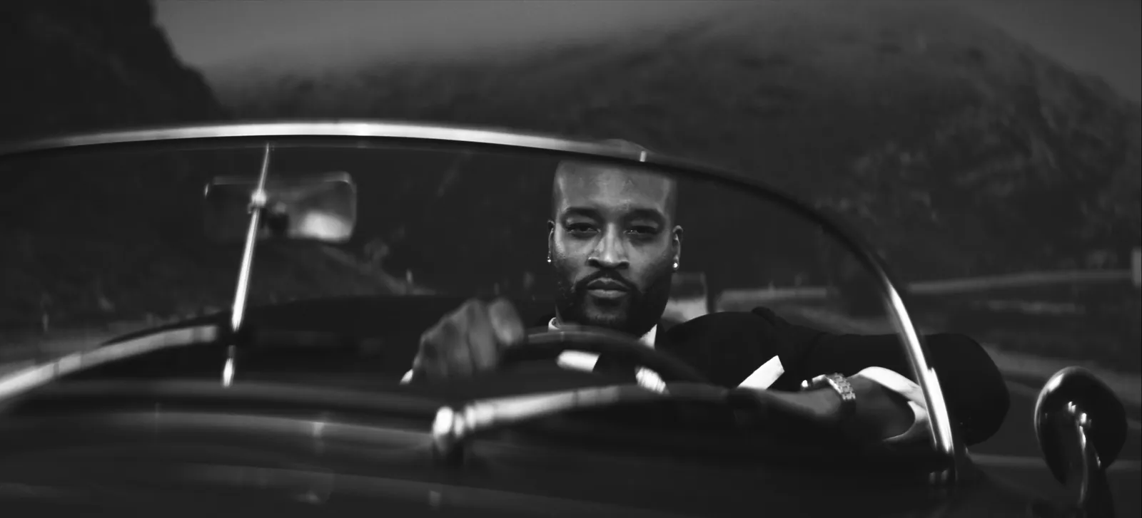 Black-and-white film still of a suited man at the wheel of a convertible.