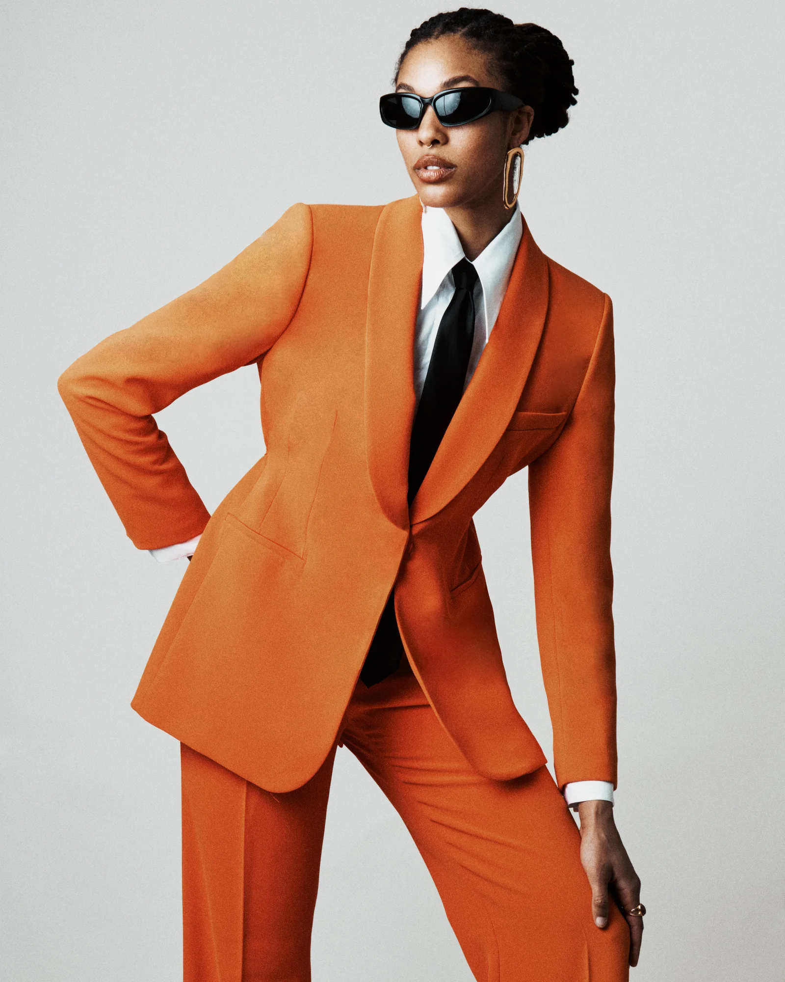 A model in orange tailoring, a black tie and sunglasses against a pale studio background.