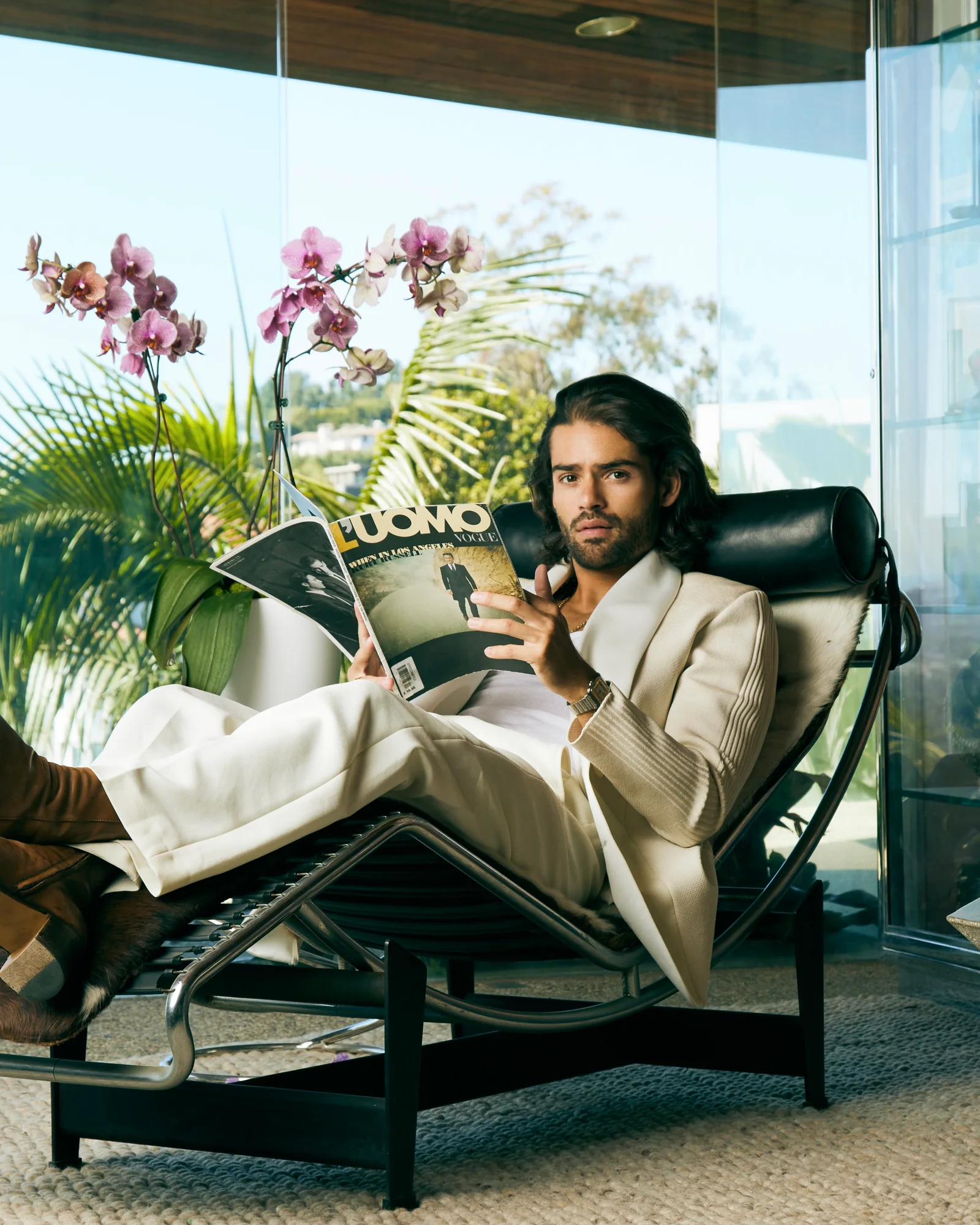 A model reading a magazine in a reclining chair beside large windows.
