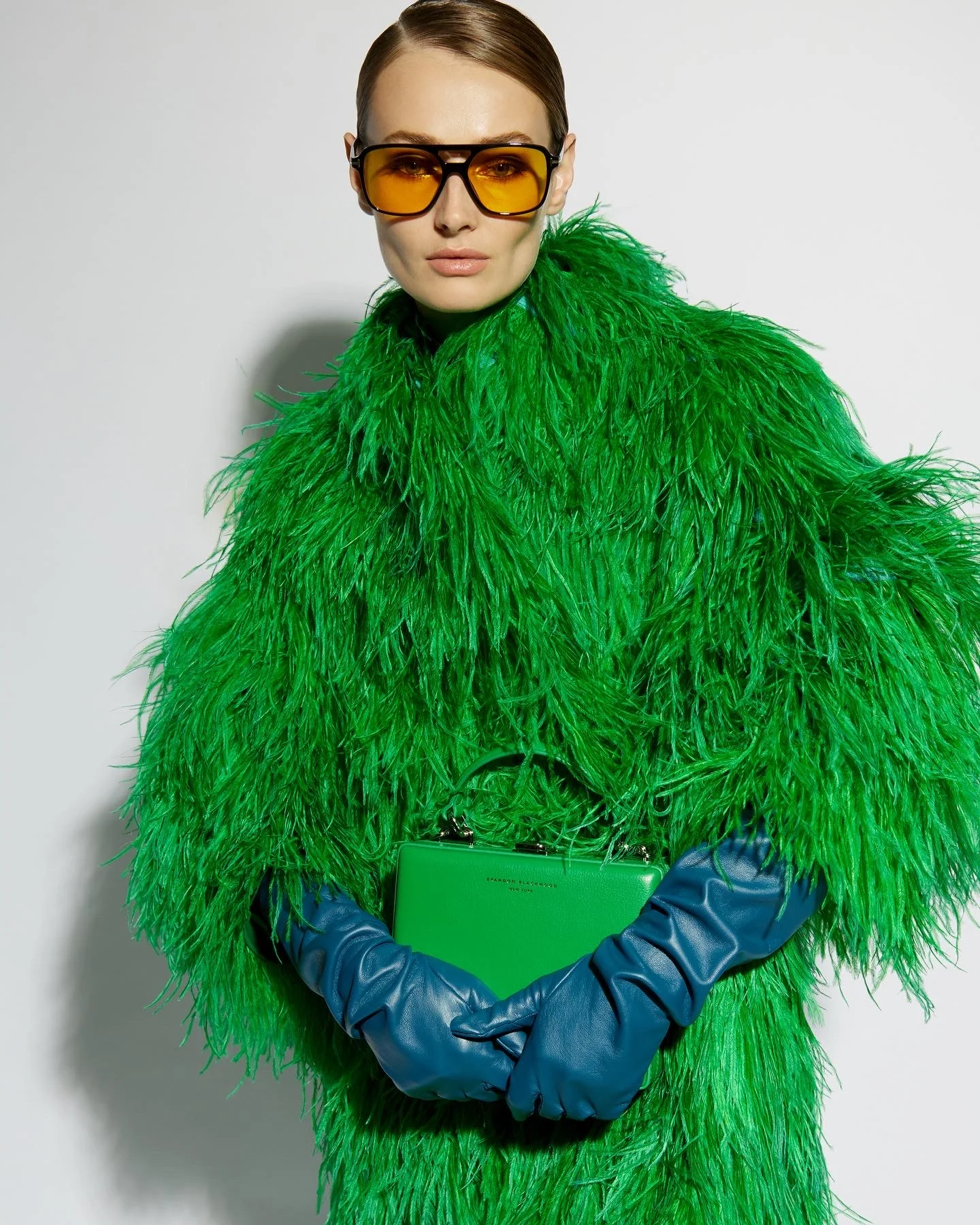 A model in a green feather coat, yellow sunglasses and blue gloves holding a green bag.
