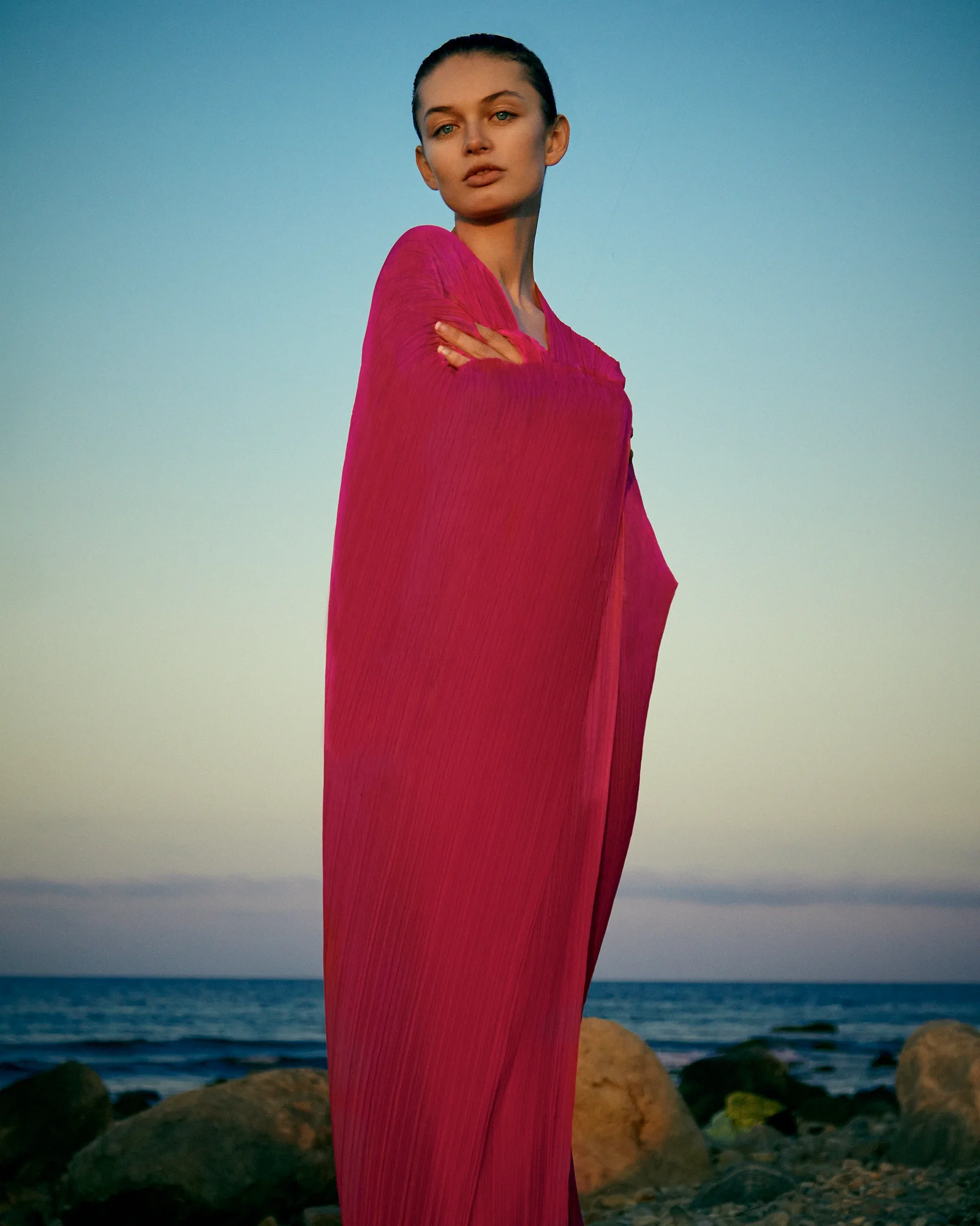 A model in pink chiffon standing by the water beneath a blue sky.
