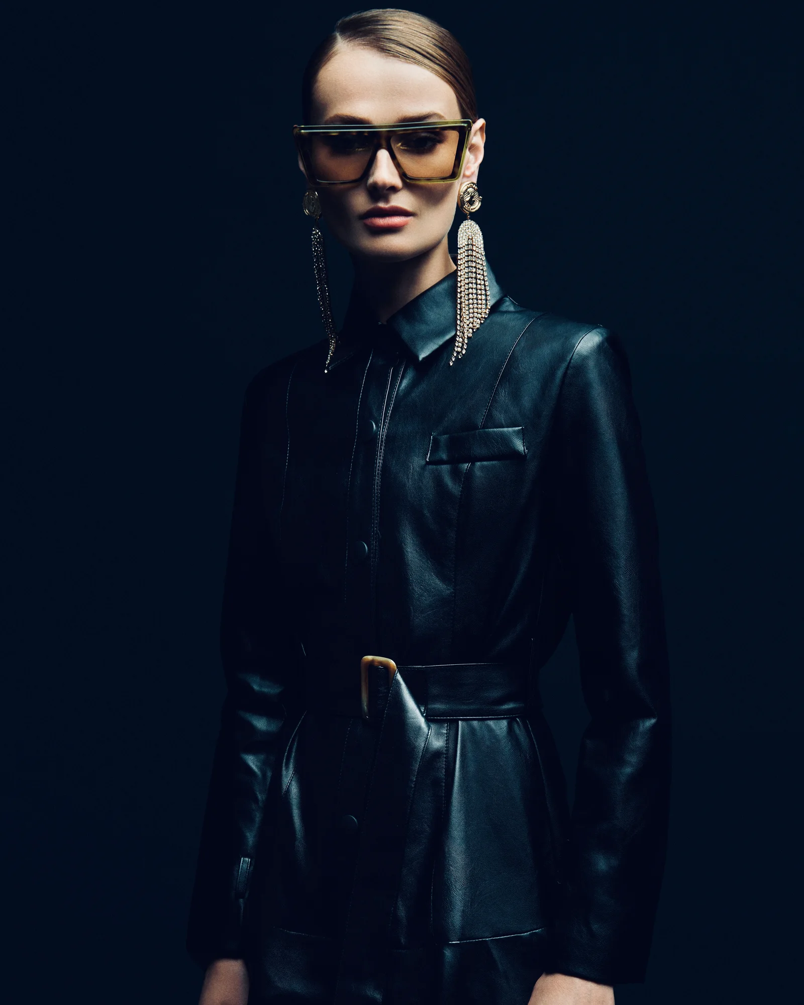 A model in dark leather, sunglasses and long earrings against a deep blue background.